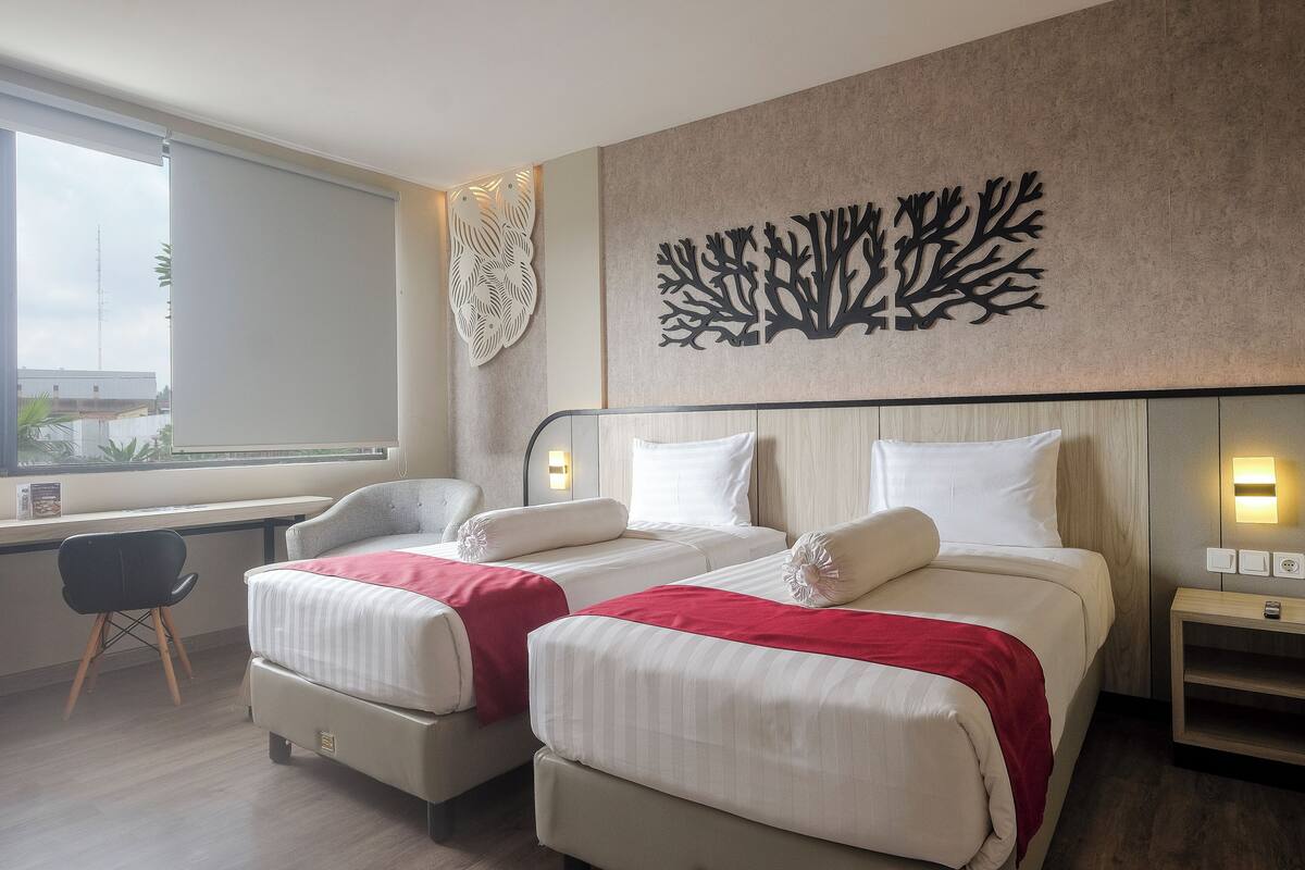 A hotel room with two beds, a desk, and a window with a view of buildings.