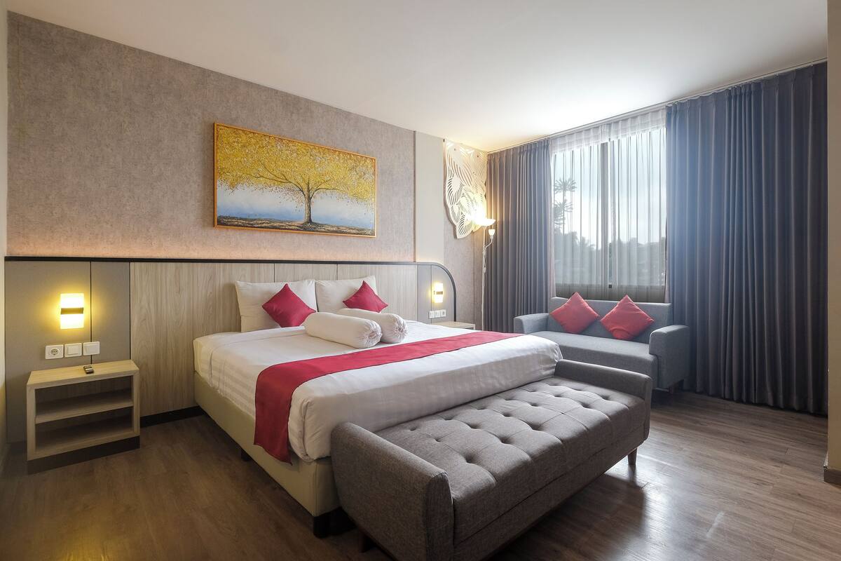 A modern hotel room with a large bed, a grey sofa, a wooden nightstand, and a painting of a tree on the wall.