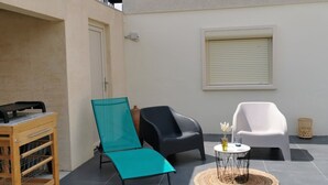 An outdoor patio with a wooden cart, a teal lounge chair, a black lounge chair, a white chair, and a small round table with a woven mat. - Quiet studio with terrace (Rougemont)