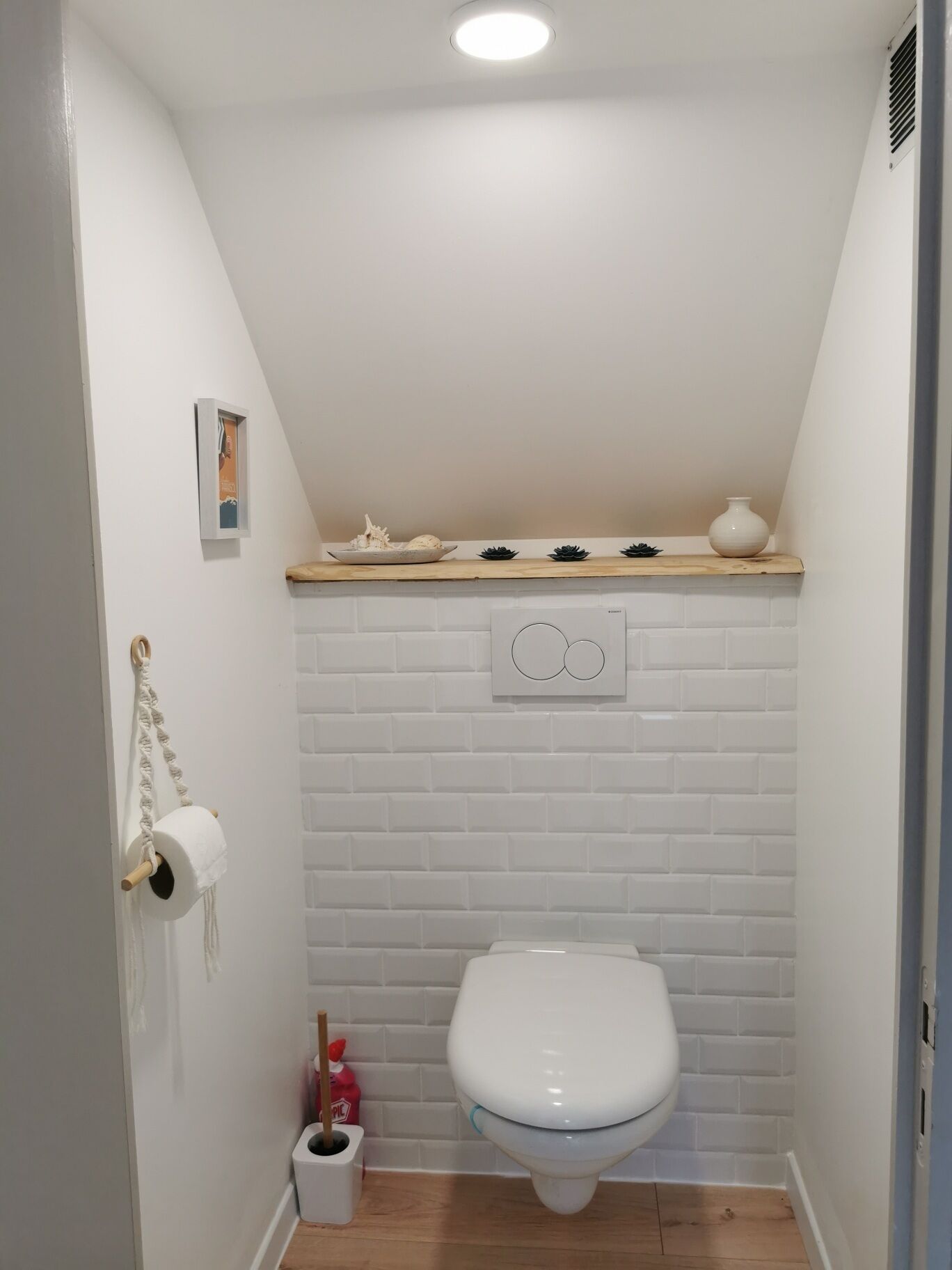 Une petite salle de bain aux murs recouverts de carreaux blancs, comprenant des toilettes, une étagère avec des objets décoratifs, un porte-rouleau de papier toilette et une petite poubelle.