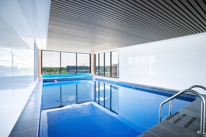 An indoor swimming pool with clear glass walls, a view of the cityscape, and a modern ceiling design.