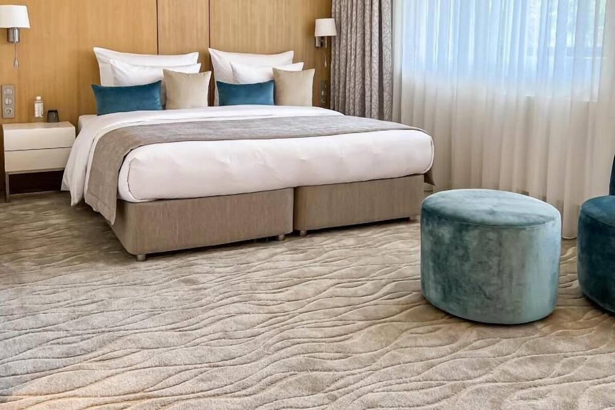 Habitación de hotel con una cama grande, dos mesitas de noche, un pouf azul y una ventana con cortinas.