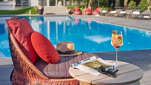 A poolside lounge with a wicker chair, a red cushion, a straw hat, a glass of drink, and a magazine. - Normafa Hotel (Budapest)
