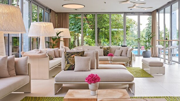 A spacious living room with a sofa set, a coffee table, and a view of a swimming pool through large windows. - Normafa Hotel (Budapest)