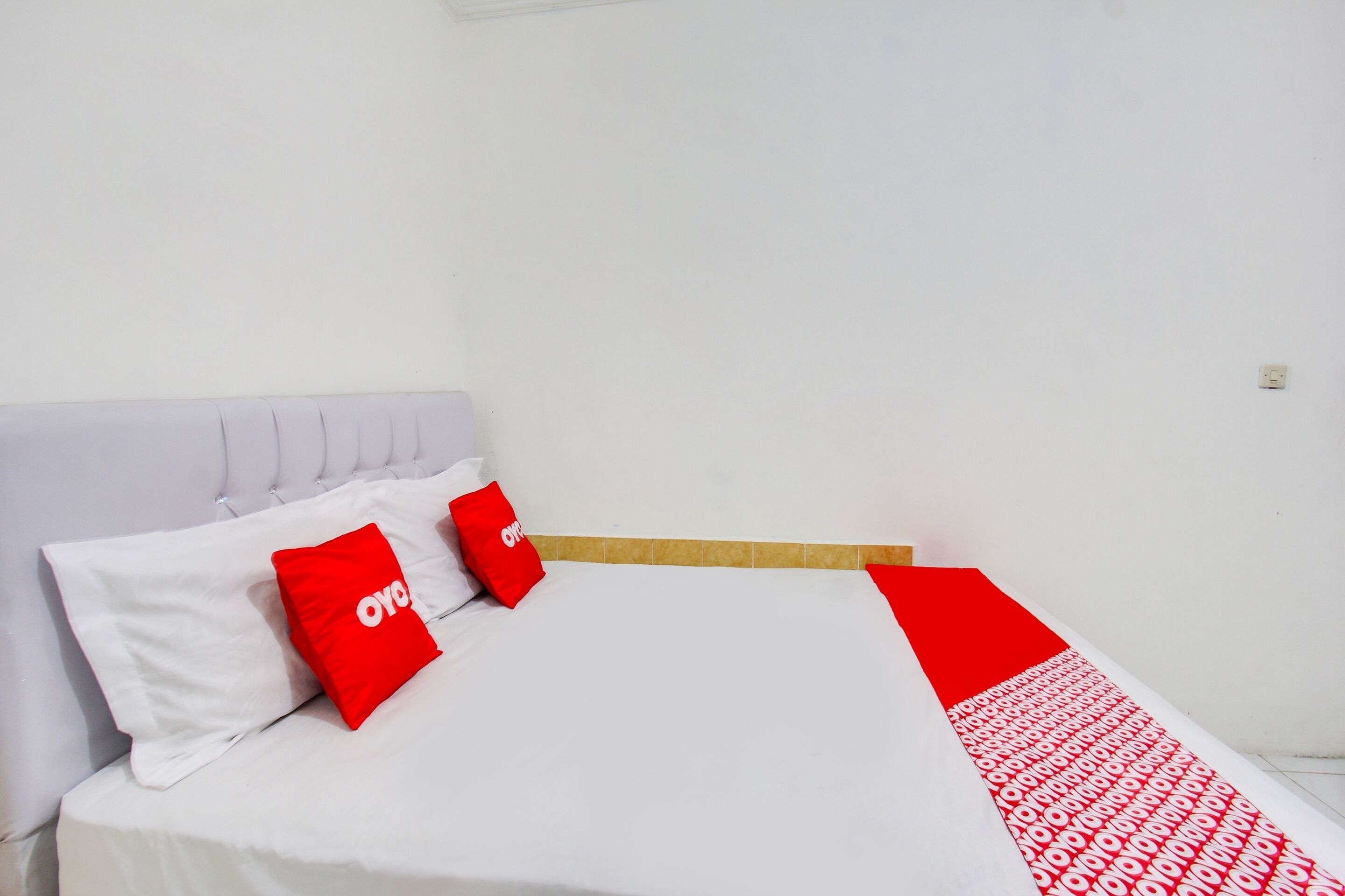 A neatly made bed with white and red bedding, a wooden headboard, and a plain white wall.