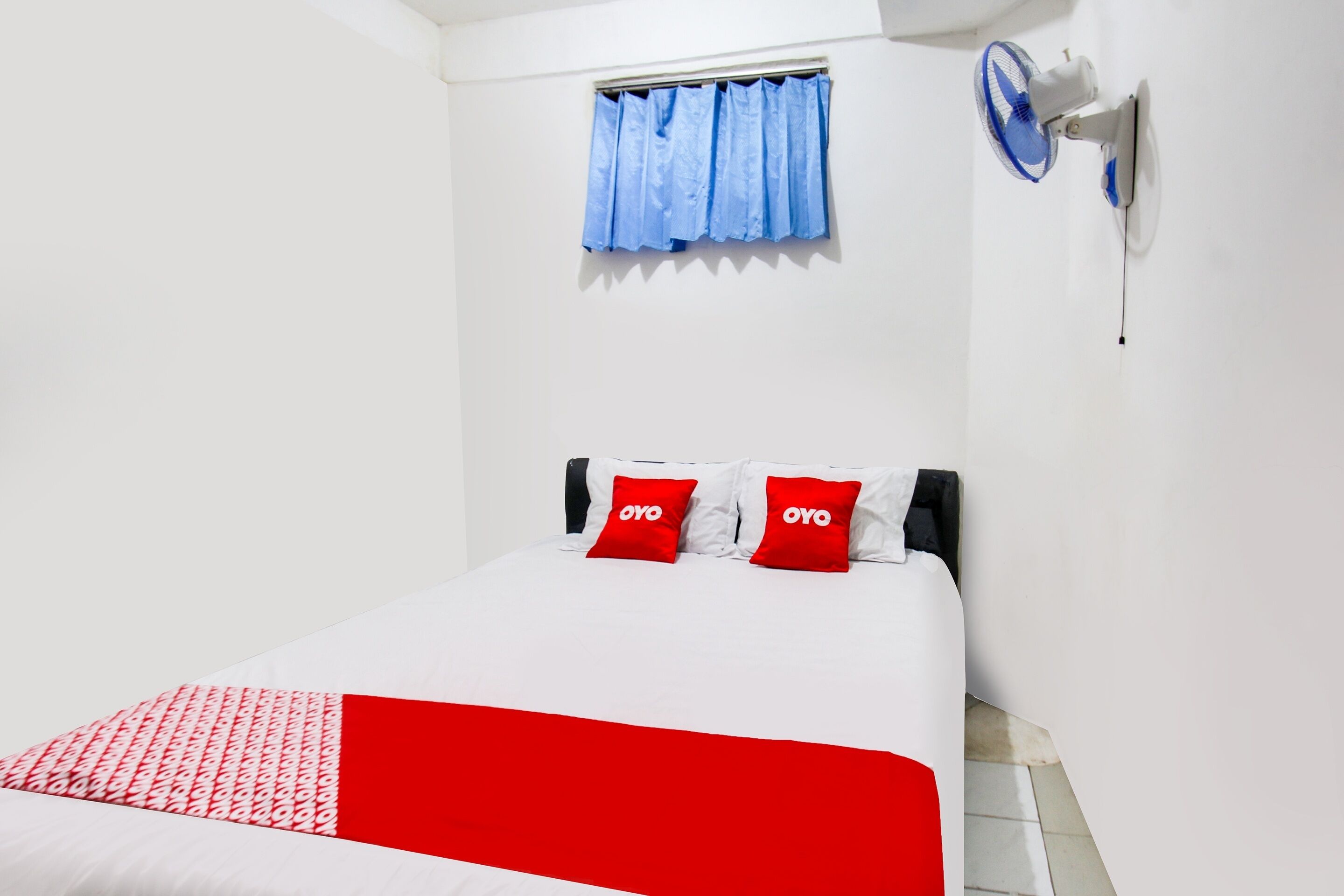 A hotel room with a bed, two red pillows, a white bedspread, a blue curtain, and a wall-mounted fan.
