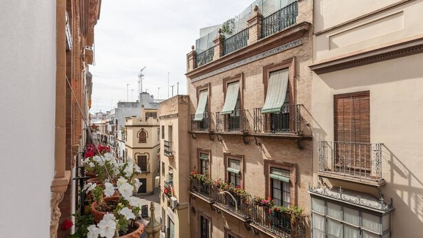 Una stretta via cittadina con edifici a più piani, balconi fioriti e cielo sereno.
