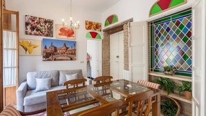 Zona pranzo con tavolo in vetro e sedie di legno, un divano grigio e una parete con opere d'arte colorate e una finestra a vetri policromi.
