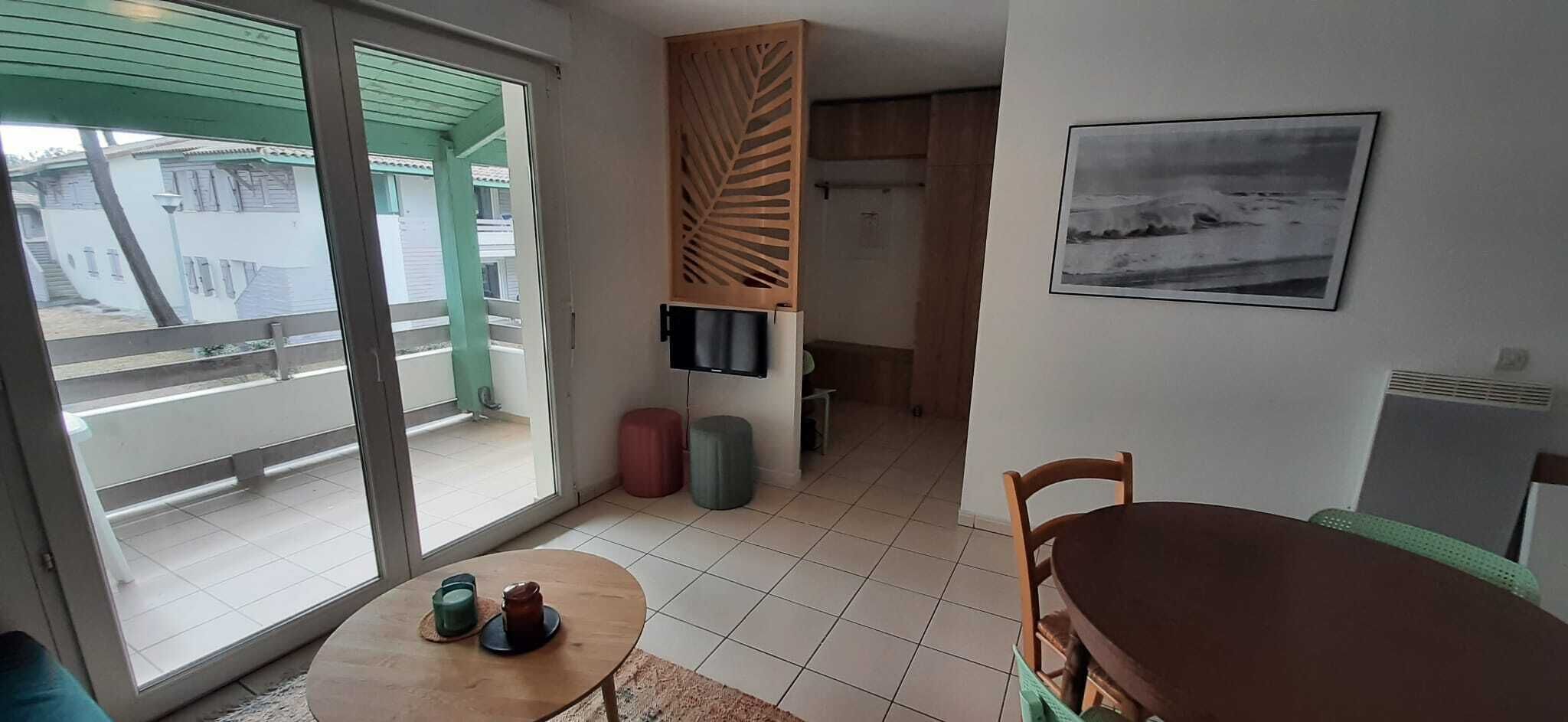 A room with a sliding glass door leading to a balcony, a small round table with a few items, a wooden chair, a television mounted on the wall, and a framed picture.