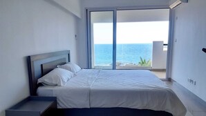 A modern bedroom with a large bed, a nightstand, and a view of the ocean through a sliding door. - La Falaise Seafront Villa (Flic En Flac)