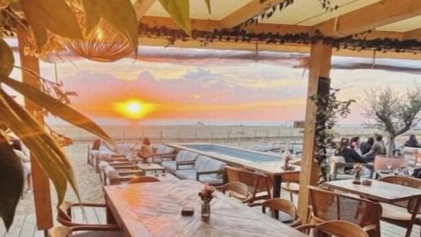 Un área de comedor al aire libre con una mesa y sillas de madera, con vista a un cuerpo de agua durante la puesta de sol.