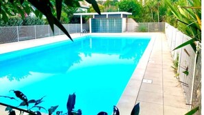 Una piscina rectangular con superficie de agua azul cristalina, rodeada por un solado de baldosas y una cerca metálica.