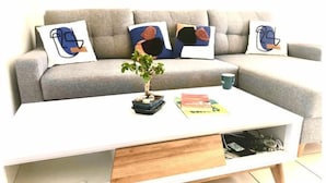 Una sala de estar moderna con un sofá gris, almohadas abstractas, una mesa de centro blanca con una planta y una revista.