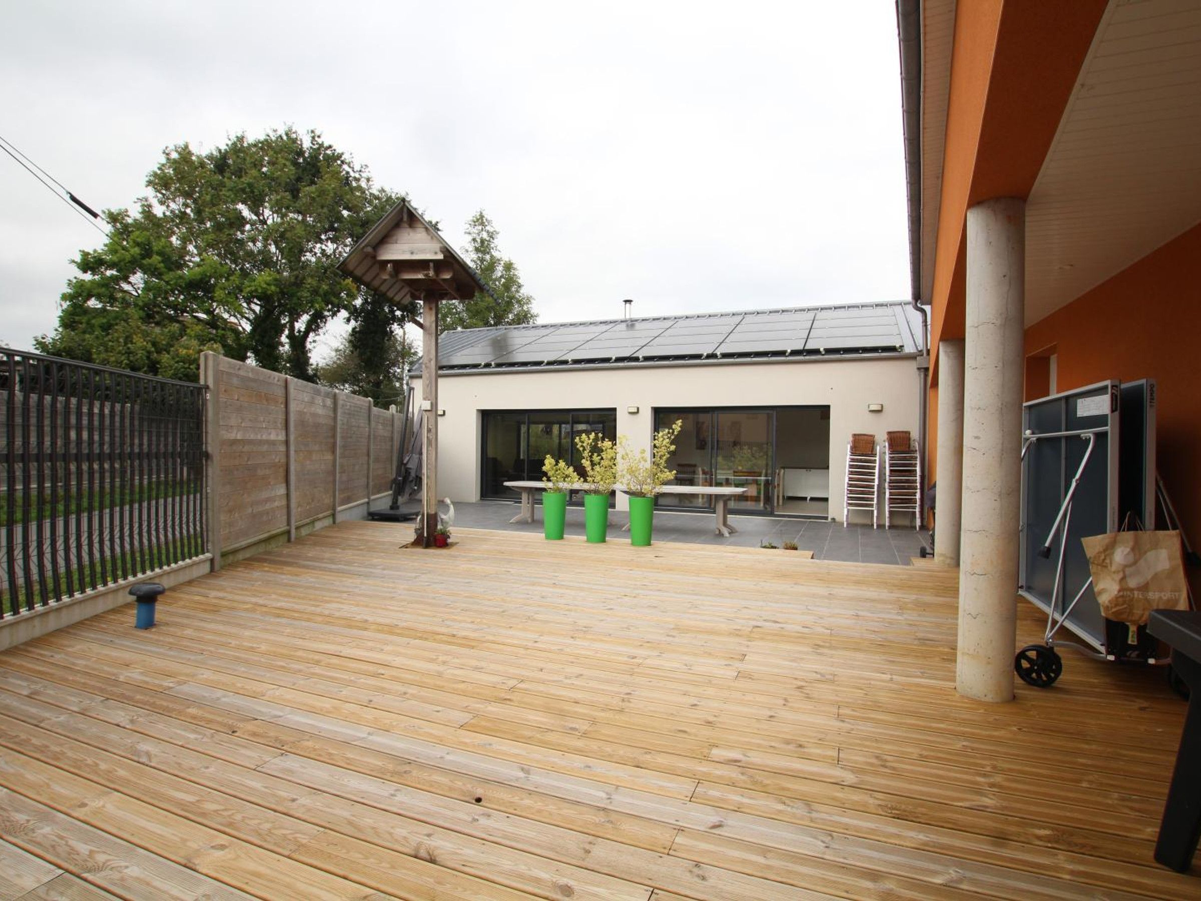 Une terrasse en bois avec une table et des chaises, une poubelle verte et une clôture en métal.