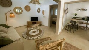 Ein Wohnzimmer mit einer Couch, einem Fernseher, einem Couchtisch und einem Korbstuhl. An den Wänden befinden sich Dekorationen und eine Pendelleuchte.