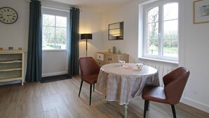 Ein Essbereich mit einem runden Tisch, zwei Stühlen, einer Pendelleuchte, einer Uhr, einem Fenster mit Vorhängen, einem Schrank und einem Bücherregal.