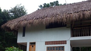 Una casa de dos pisos con techo de paja, paredes blancas y una ventana pequeña con diseño de celosía.
