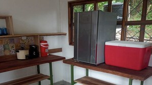 Una cocina compacta con refrigerador, hielera roja, cafetera y una olla sobre una repisa.