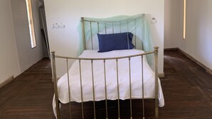 Una cama individual con estructura metálica y colchón blanco en una habitación con suelo de madera y paredes blancas.