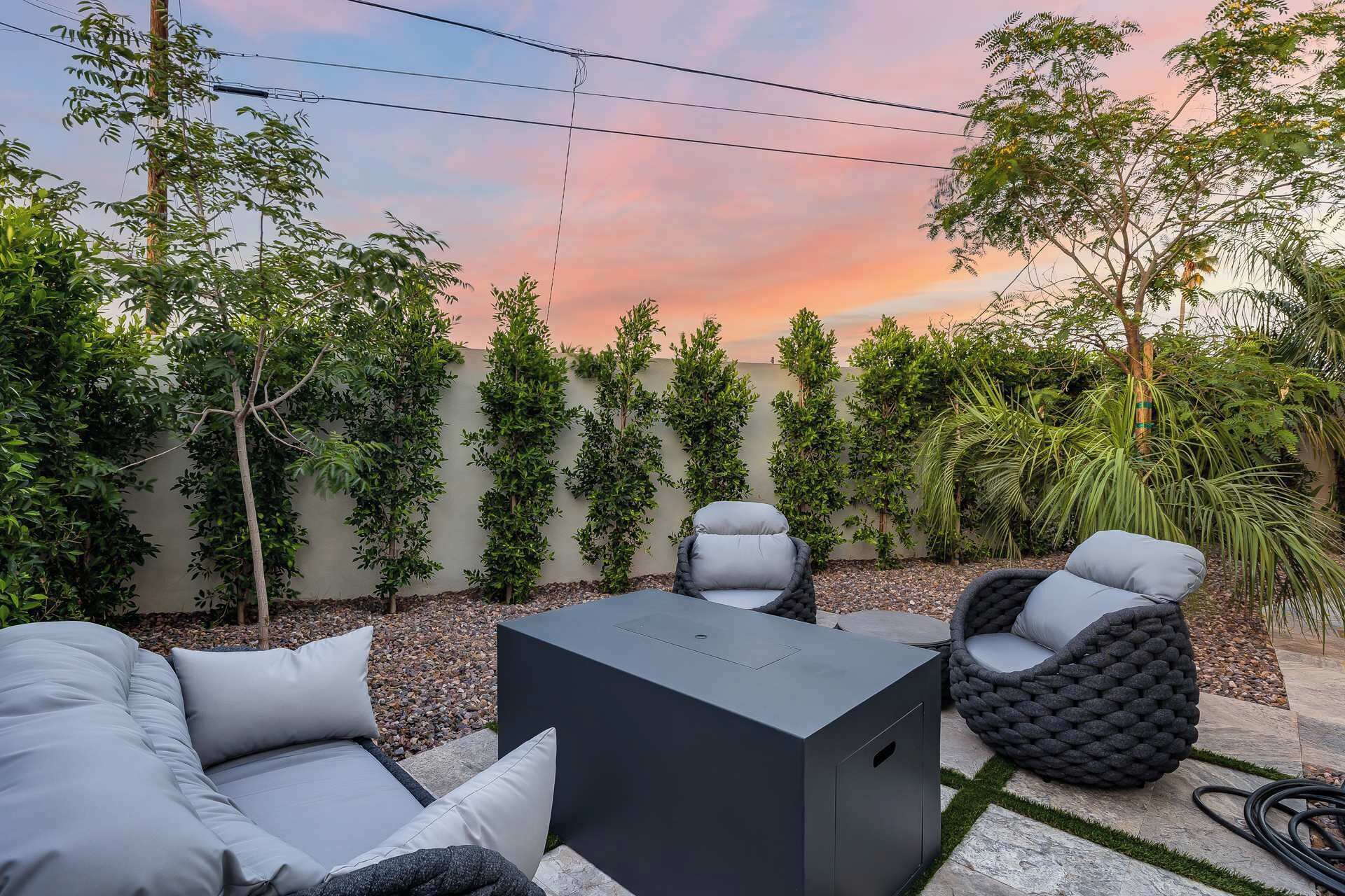 An outdoor seating area with a sofa, chairs, and a table, surrounded by a wall of greenery and a sunset sky.