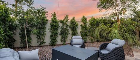 An outdoor seating area with a sofa, chairs, and a table, surrounded by a wall of greenery and a sunset sky.