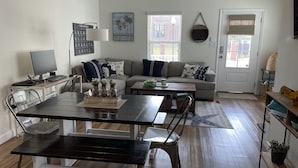 A living room with a sofa, coffee table, and a dining table with chairs. There is a TV on a stand, a lamp, and a framed alphabet chart on the wall. - Experience Downtown Charm and Modern Comfort at  Your Perfect VRBO Getaway! (Pensacola)