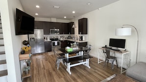 A modern kitchen with dark cabinets, stainless steel appliances, a dining area with a black table and metal chairs, and a small workspace with a computer and lamp. - Experience Downtown Charm and Modern Comfort at  Your Perfect VRBO Getaway! (Pensacola)