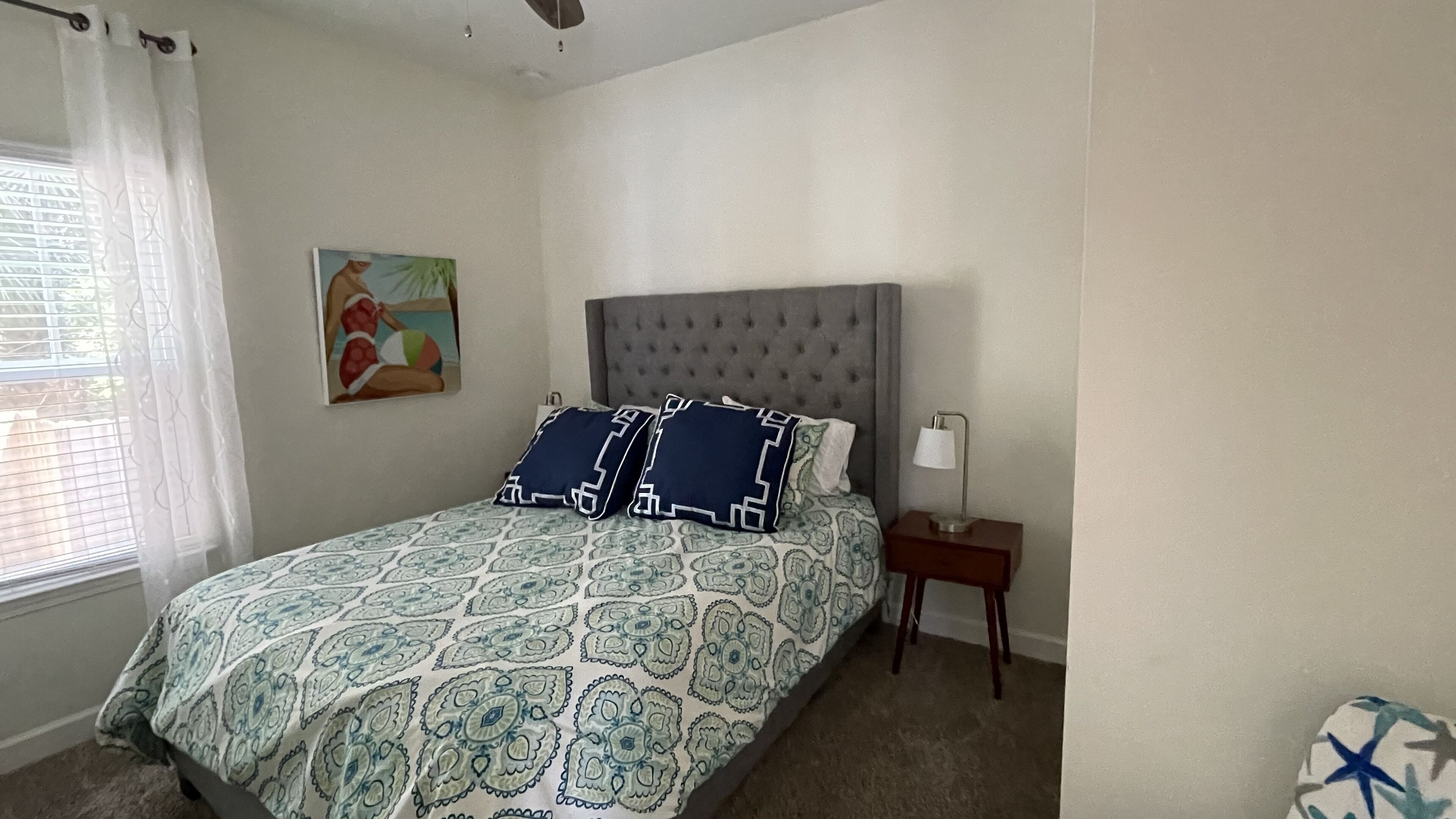Un dormitorio con cabecera tapizada, colcha estampada, dos almohadas, una lámpara de noche y un cuadro en la pared.
