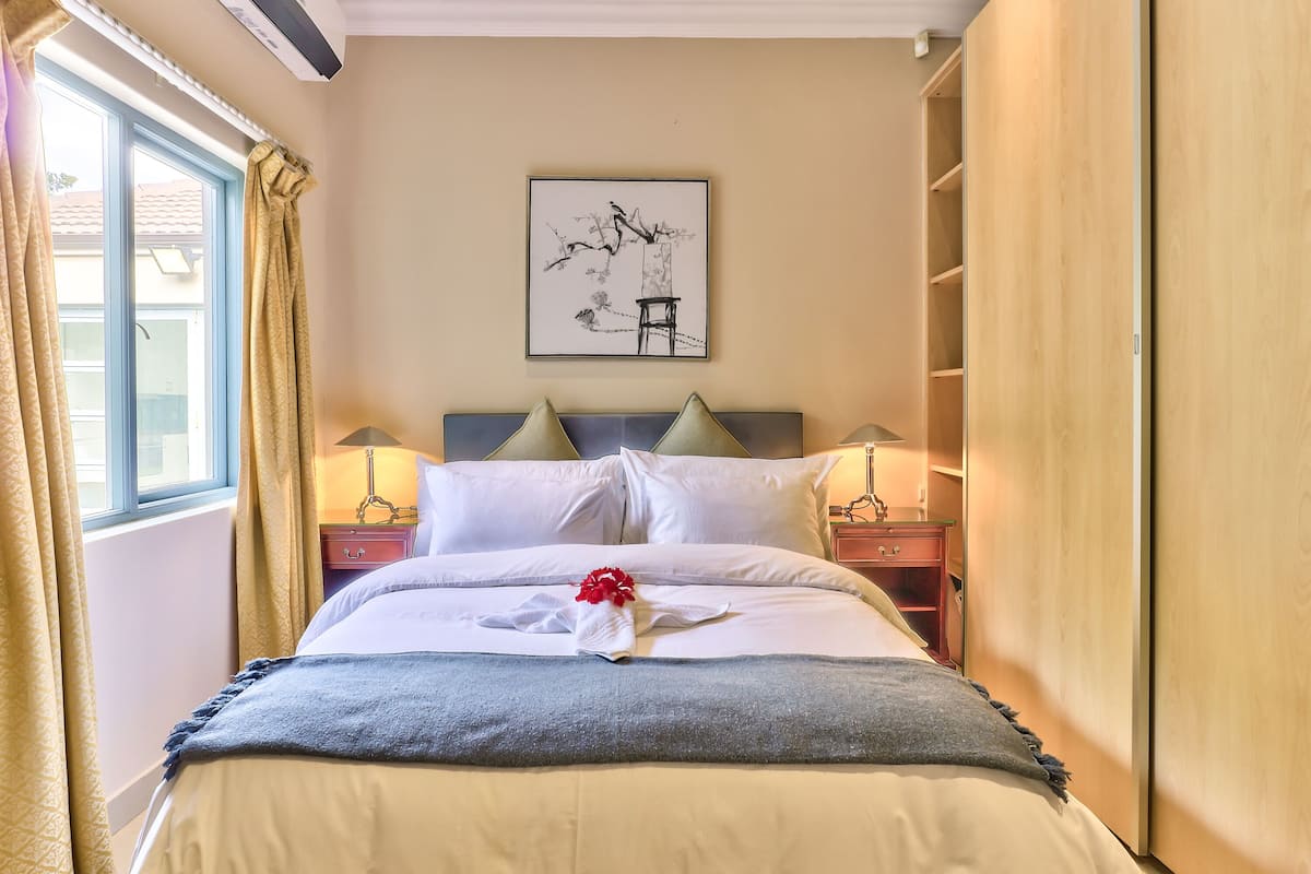 A neatly made bed with white linens and a grey blanket, a bedside table with a lamp, and a wall-mounted artwork.