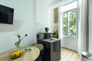 A compact kitchen with a dining area, a TV mounted on the wall, and a window with a view of greenery. - Charming Studio Apartment with kitchen (Tel Aviv)