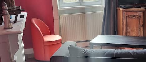 Una habitación con un ventanal grande, pared roja, sofá negro, cómoda blanca y televisor sobre un mueble de madera.