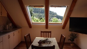A dining area with a table set for two, a window with a view of a forested hillside, and a flat-screen TV mounted on the wall. - Apartment Lile (Dubrovnik)