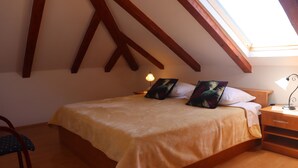 A bedroom with a sloped ceiling, wooden beams, a bed with a beige bedspread, two pillows, a nightstand with a lamp, and a chair. - Apartment Lile (Dubrovnik)