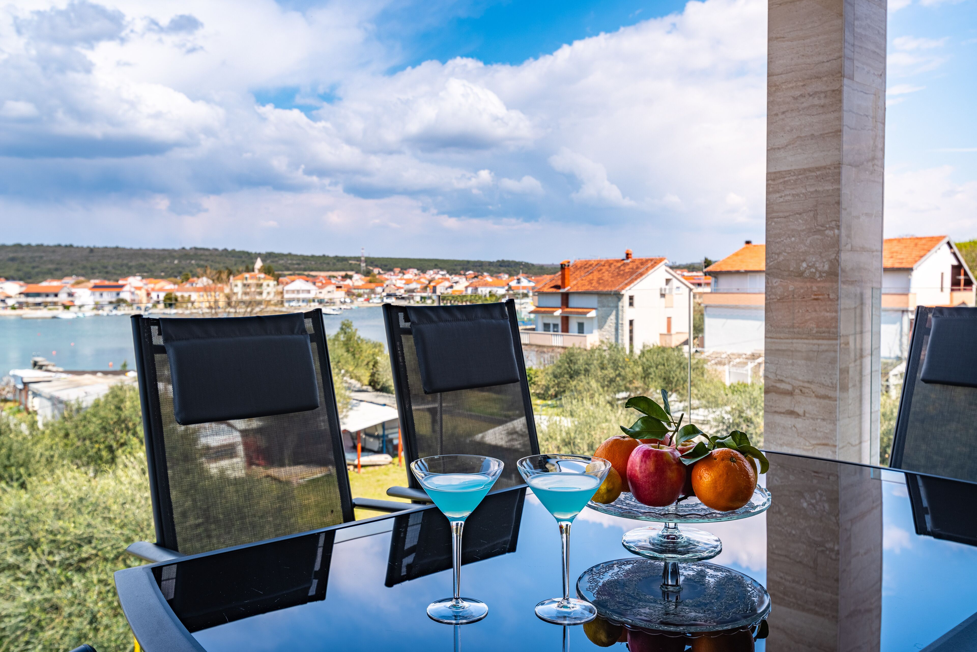 A table with two martini glasses, a fruit platter, and a view of a coastal town.