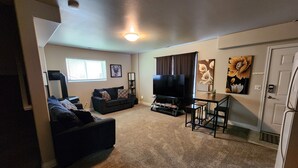 A living room with a dark-colored sofa, a flat-screen TV, a coffee table, and a dining table with chairs. There is a window with blinds, a painting on the wall, and a door to the right.