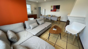 Ein modernes Wohnzimmer mit einem grauen Ecksofa, einem Holztisch und einem weißen Sessel. Im Hintergrund befindet sich ein Essbereich mit Tisch und Stühlen.
