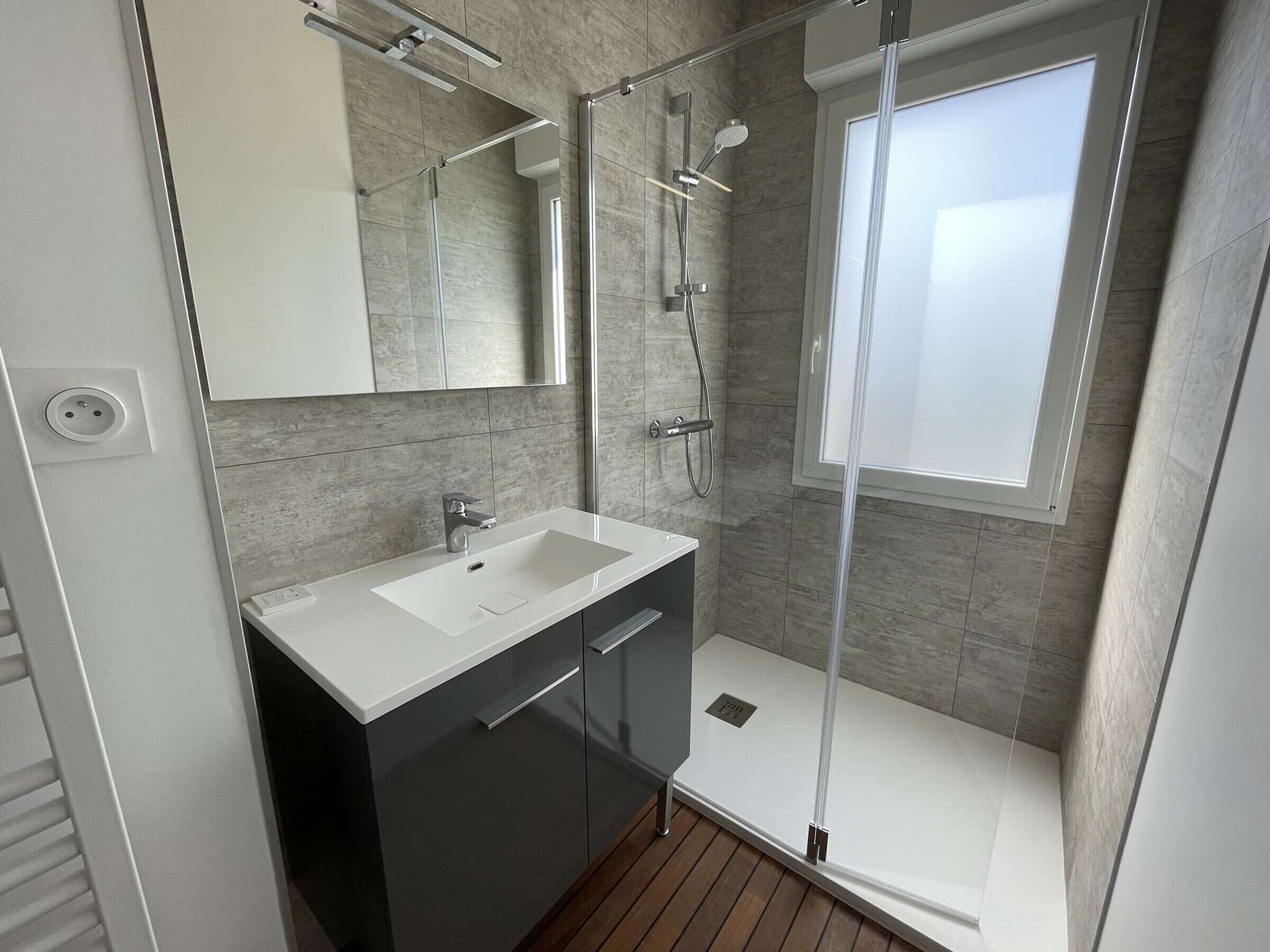 A modern bathroom with a white sink, dark cabinets, a glass-enclosed shower, and a large window.