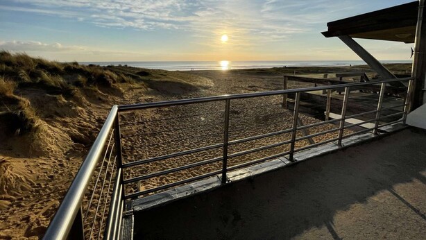Ein Strand mit einer Metalltreppe, die zum Sand führt, einer Holzpromenade und einem klaren Himmel, in dem die Sonne untergeht.