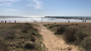 Ein Strand mit einem sandigen Pfad zum Wasser, einem Steg, der ins Meer führt, und Menschen, die am Ufer entlanggehen.