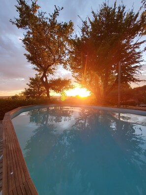 Una piscina con cubierta de madera, rodeada de árboles y un atardecer al fondo.