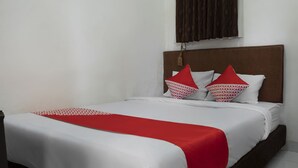 Ein ordentlich bezogenes Bett mit weißer und roter Bettwäsche, ein braunes Kopfteil und ein Fenster mit gemusterten Vorhängen.