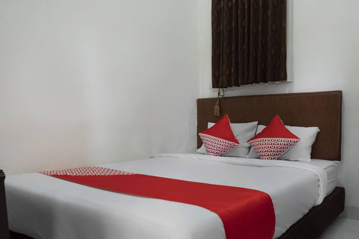 A neatly made bed with white and red bedding, a brown headboard, and a window with patterned curtains.