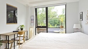 A bedroom with a large bed, a desk with a chair, a potted plant, and a view of greenery through a sliding door. - Wandle Waters, by Veeve (London)