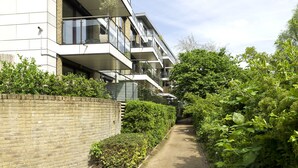 A modern apartment building with multiple balconies, a brick wall, and a pathway surrounded by greenery. - Wandle Waters, by Veeve (London)