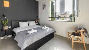 Ein modernes Schlafzimmer mit einem großen Bett, einem Beistelltisch mit Pflanze, einem Sessel mit Laptop und einem Fenster mit Blick auf die Stadt.