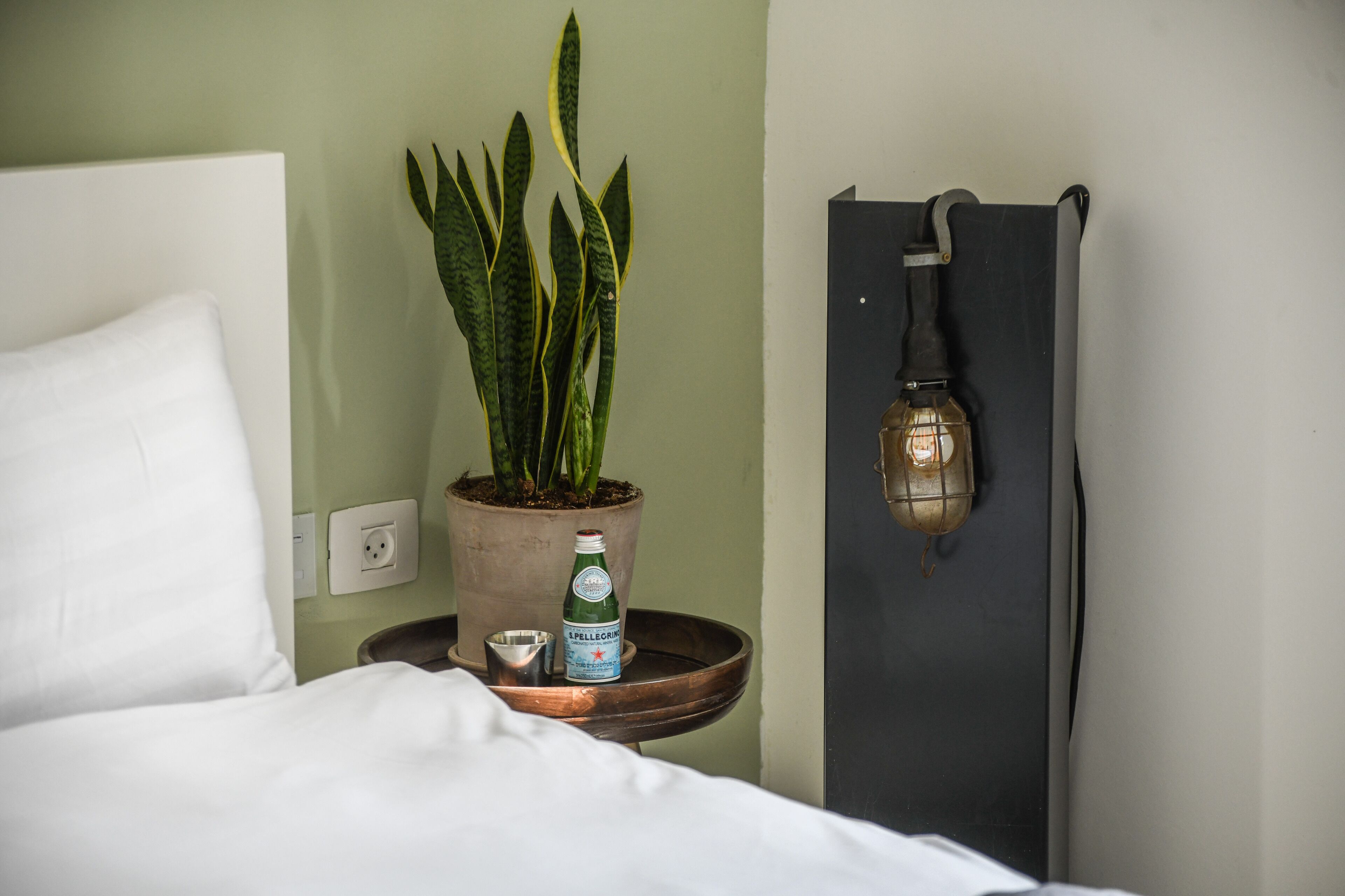 A neatly made bed with white linens, a potted plant, a bottle of L'Pellegrino water, and a wall-mounted light fixture.