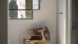 Ein hölzerner Stuhl mit einer schwarzen Tasche und einem grauen Schal, vor einem Fenster mit Blick auf Gebäude und einen Baum.