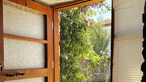 An open sliding glass door leading to a garden with a stone wall, a small tree, and a tiled floor. - Vasvija Pansiyon (Ayvalik)