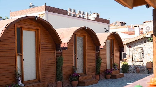 A row of wooden arched rooms with doors, potted plants, and a wooden table with a kettle and cup. - Vasvija Pansiyon (Ayvalik)