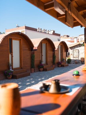 A row of wooden arched rooms with doors, potted plants, and a wooden table with a kettle and cup. - Vasvija Pansiyon (Ayvalik)